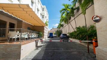 Uma rua estreita com portão, palmeiras e um edifício com fachada branca e área de estar coberta ao ar livre.