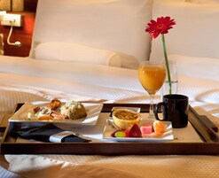 Bandeja con alimentos para el desayuno, como bollería, frutas y un vaso de jugo de naranja, sobre una cama con ropa blanca.