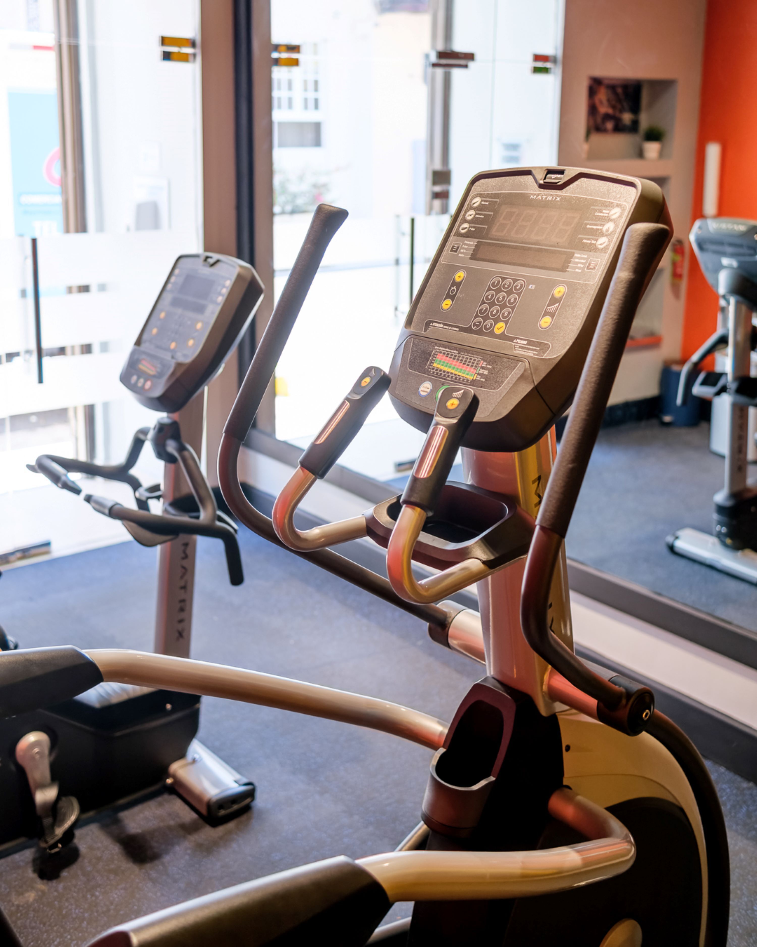 An exercise machine with a digital display and control panel, positioned in a gym with a window view.