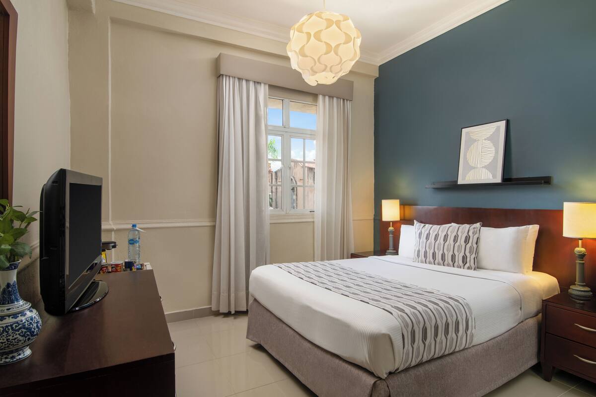 Ein Hotelzimmer mit Bett, Nachttischen, Fernseher, Fenster mit Vorhängen und einem Wandbild.