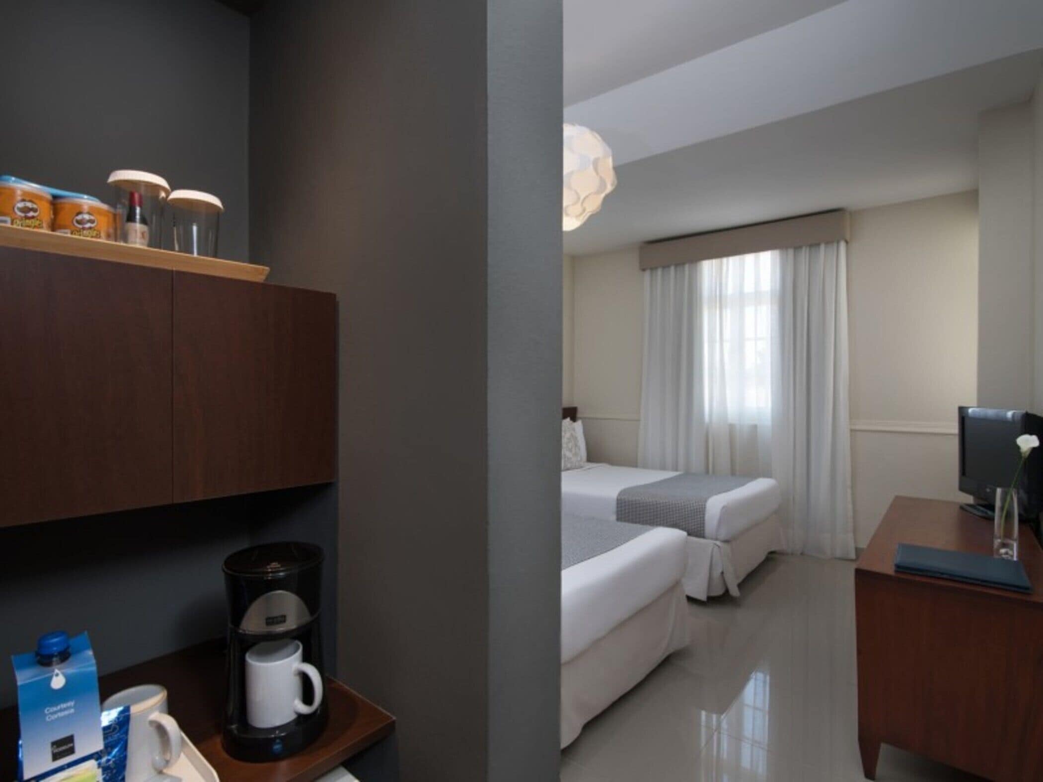 Habitación de hotel con cama, escritorio con computadora, cafetera y repisa con artículos de aseo.