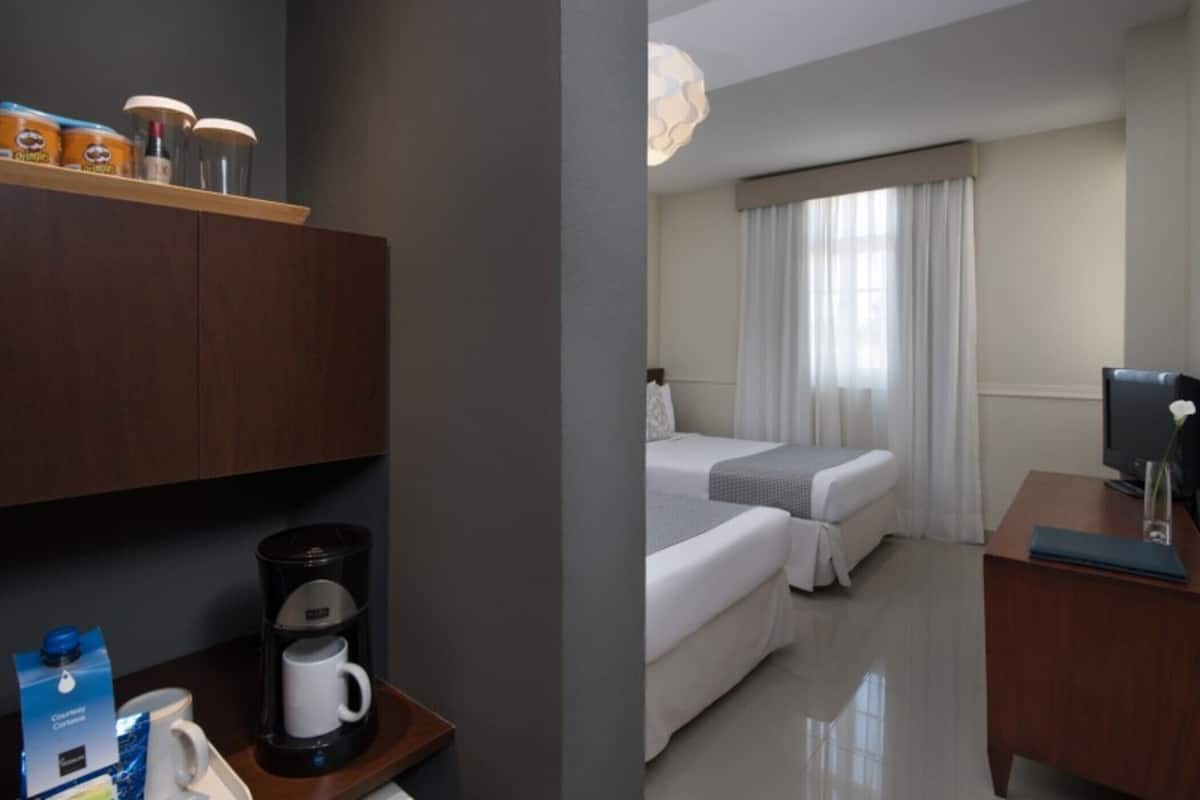 Habitación de hotel con cama, escritorio con computadora, cafetera y repisa con artículos de aseo.
