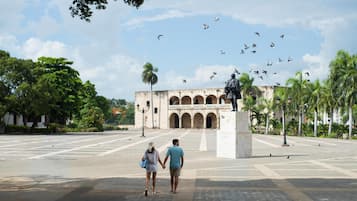 Uma praça ampla com um monumento central, rodeada por palmeiras e um edifício com arcos.