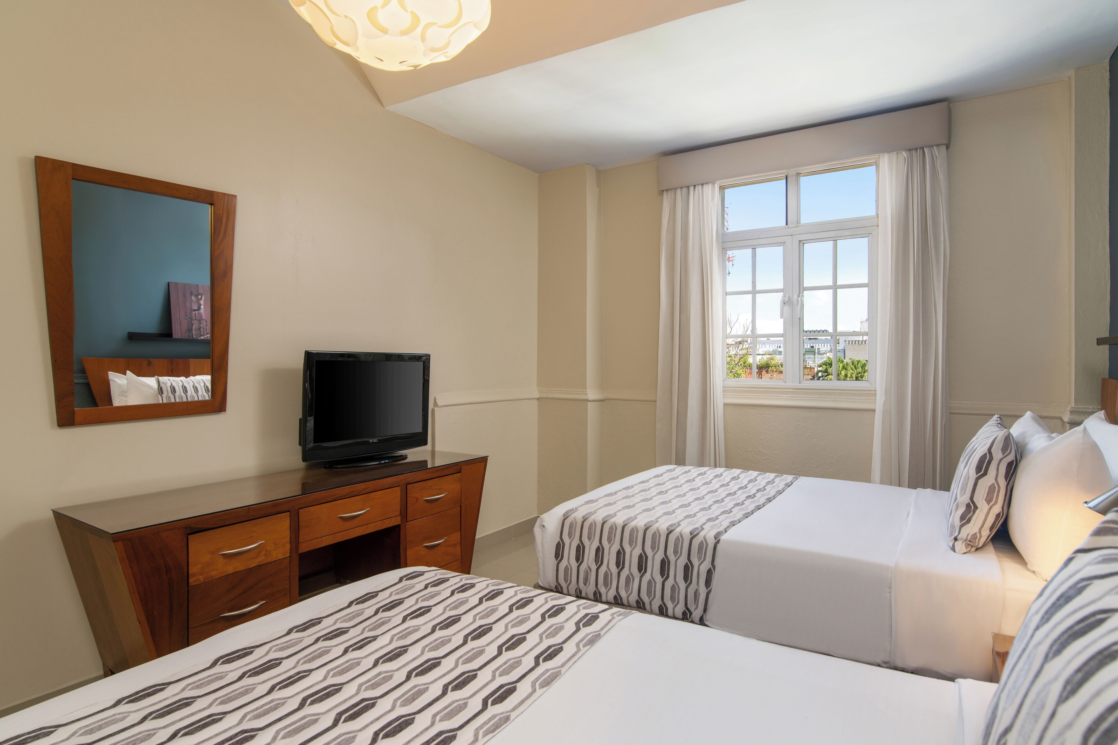 A hotel room with two beds, a TV on a wooden dresser, a large window with curtains, and a view of the city.