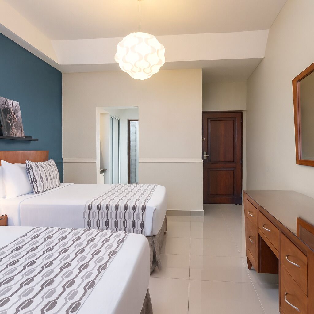 A hotel room with two beds, a wooden dresser, a hanging light fixture, and a door leading to another room.
