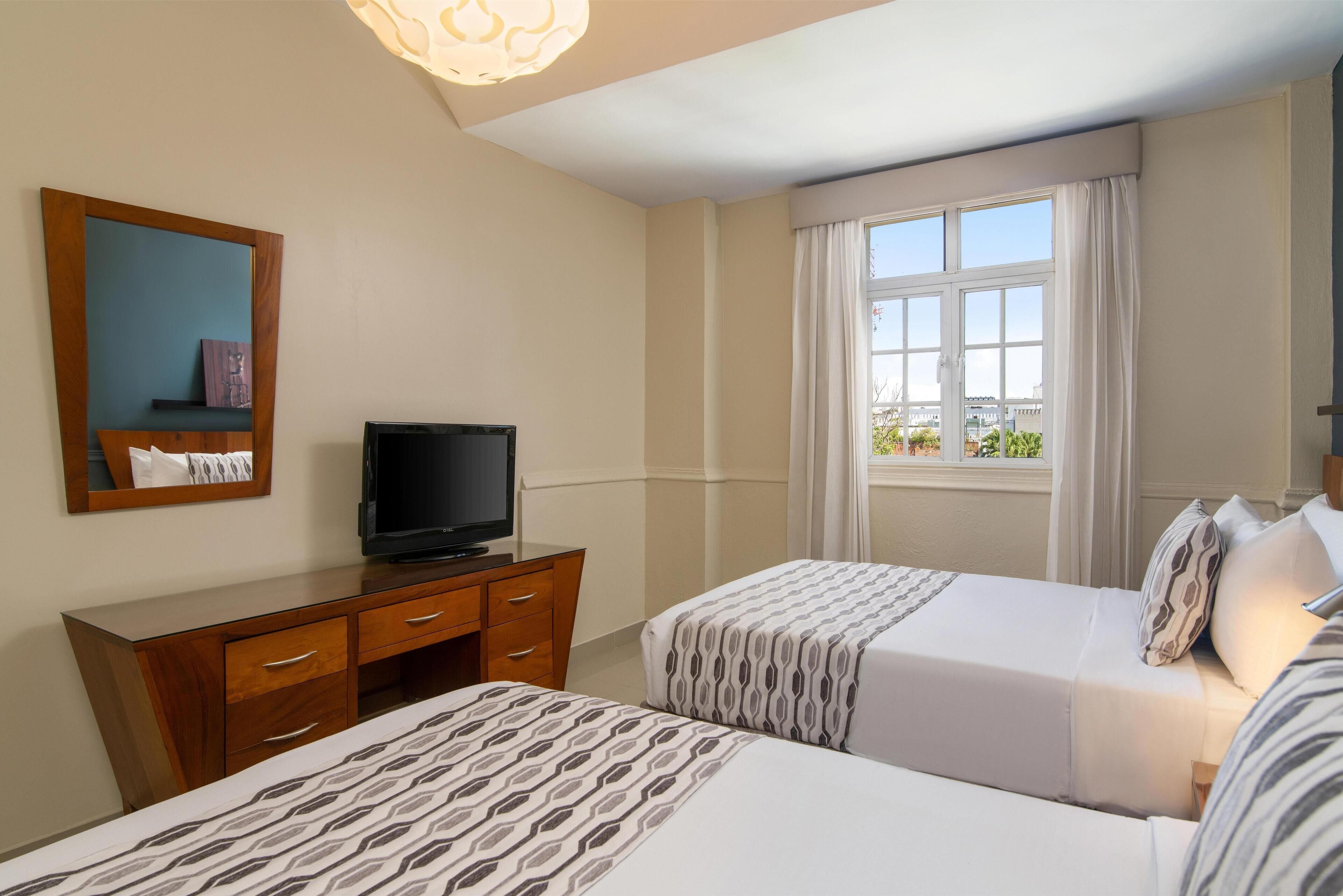A hotel room with two beds, a TV on a wooden dresser, a large window with curtains, and a view of the city.