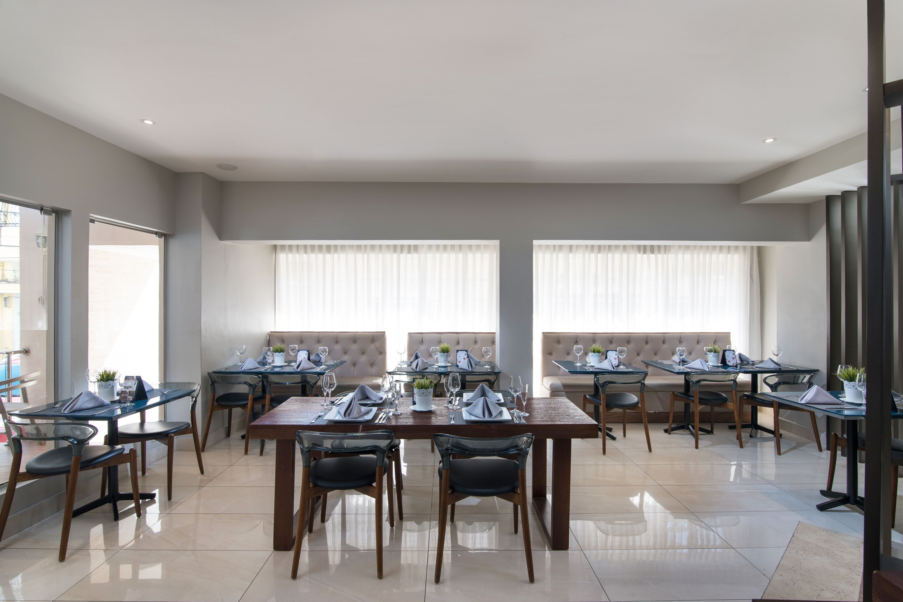 A modern dining area with multiple tables and chairs, large windows with blinds, and a clean, minimalist design.