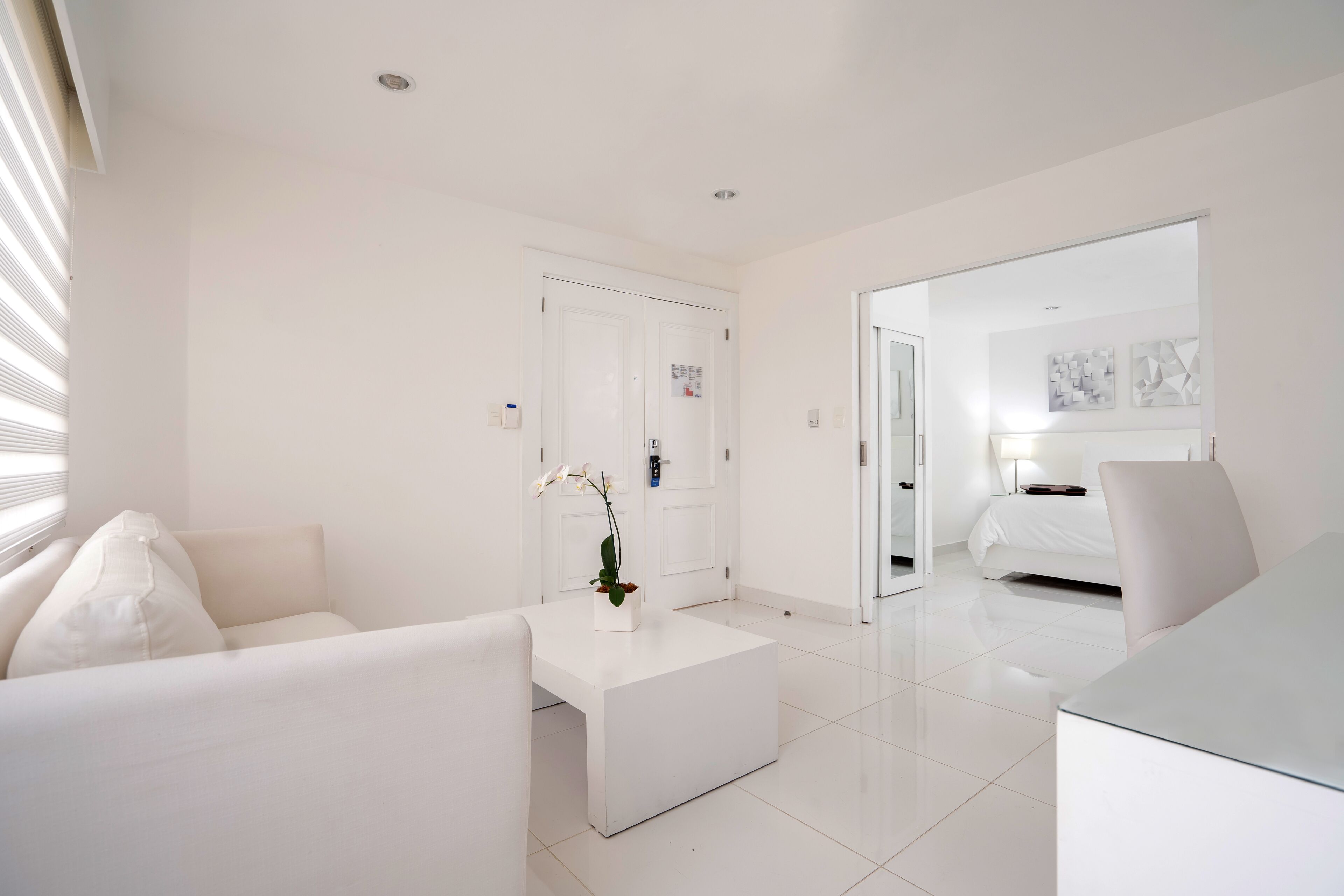 A modern hotel room with a white color scheme, a sofa, a coffee table with a plant, a bed, and a desk.
