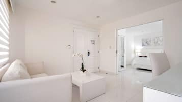 A modern hotel room with a white color scheme, a sofa, a coffee table with a plant, a bed, and a desk.