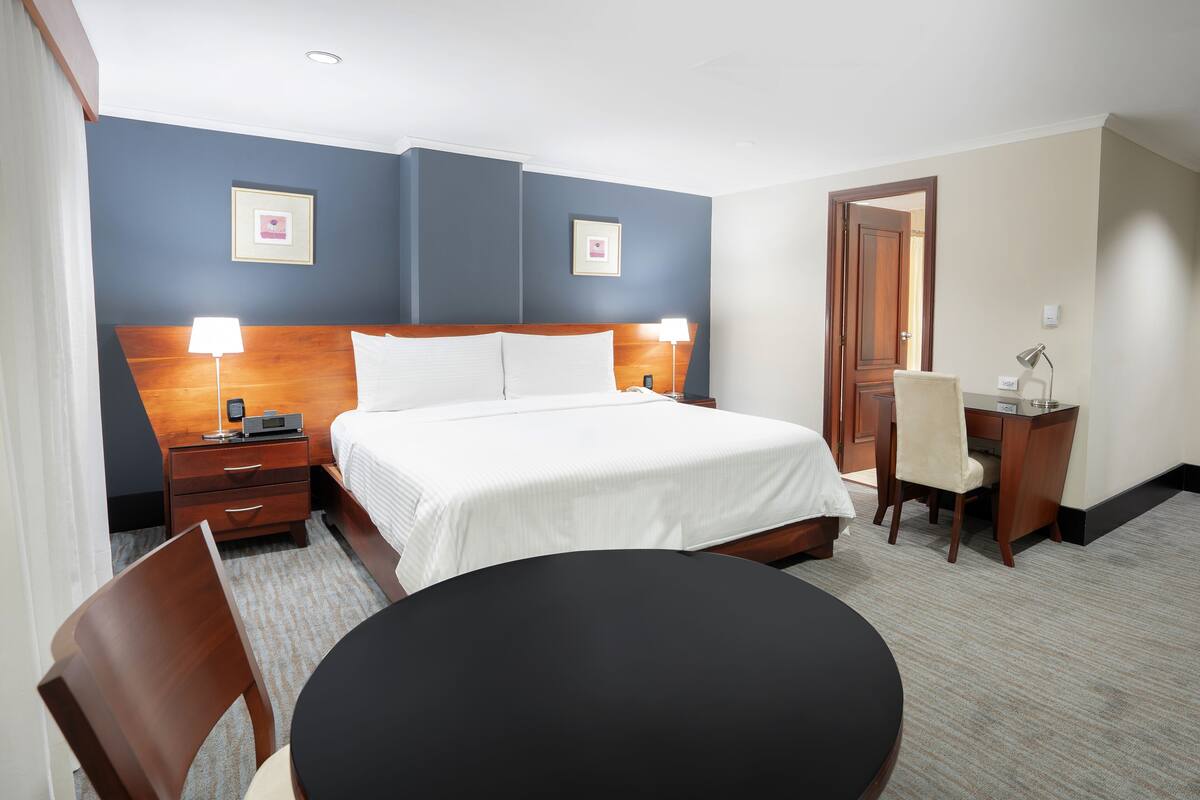 A hotel room with a bed, bedside tables, a desk, a chair, and a round table.