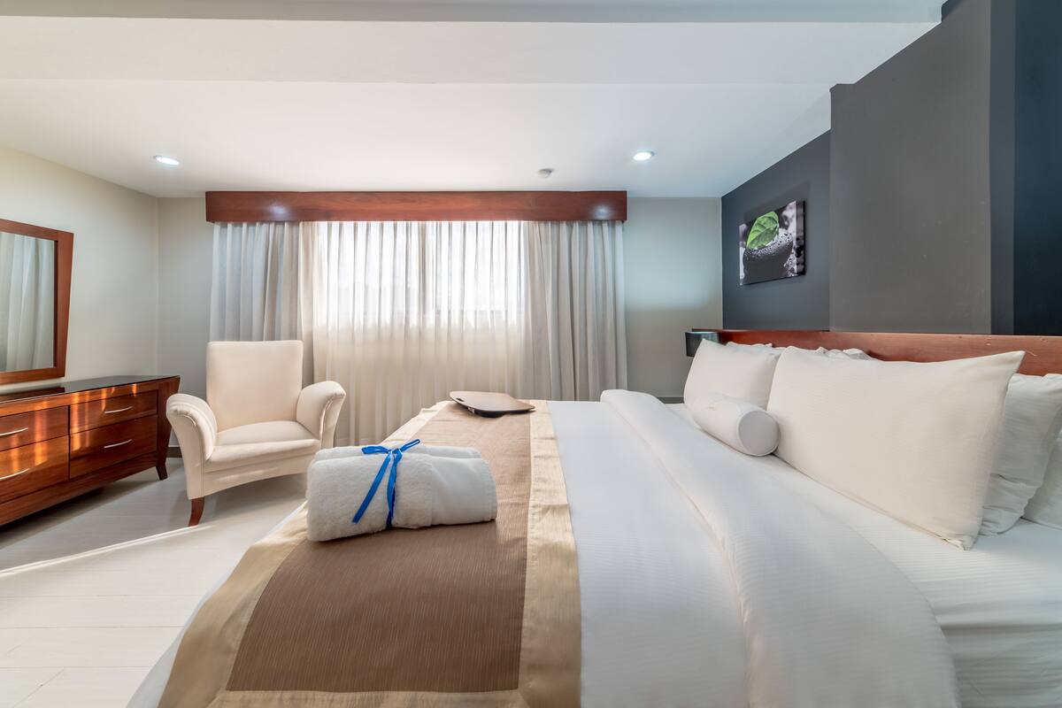 A hotel room with a bed, a chair, a dresser, a scale, and a painting on the wall.