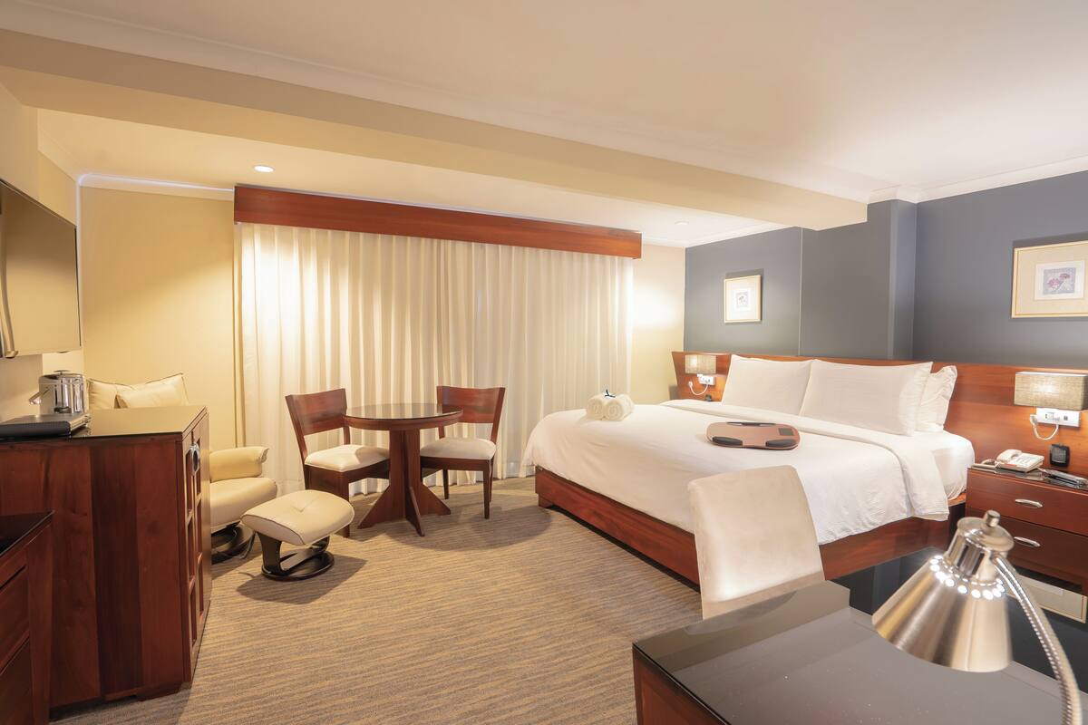 A hotel room with a bed, a desk, a chair, a table, a lamp, and a window with curtains.