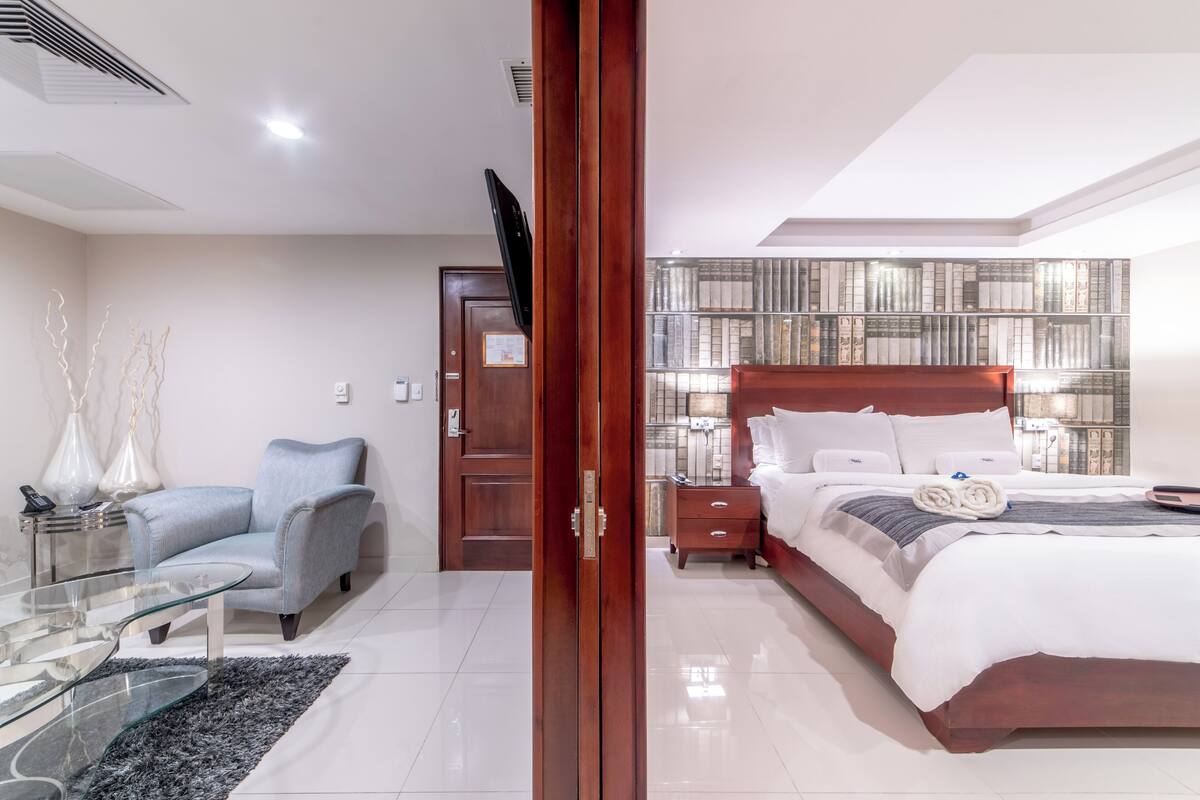 A modern bedroom with a wooden bed, a glass coffee table, a grey armchair, and a bookshelf.