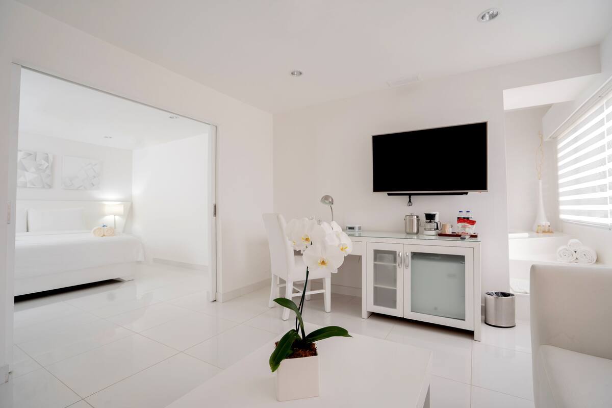 A modern hotel room with a white color scheme, a flat-screen TV, a dining area, and a potted orchid.