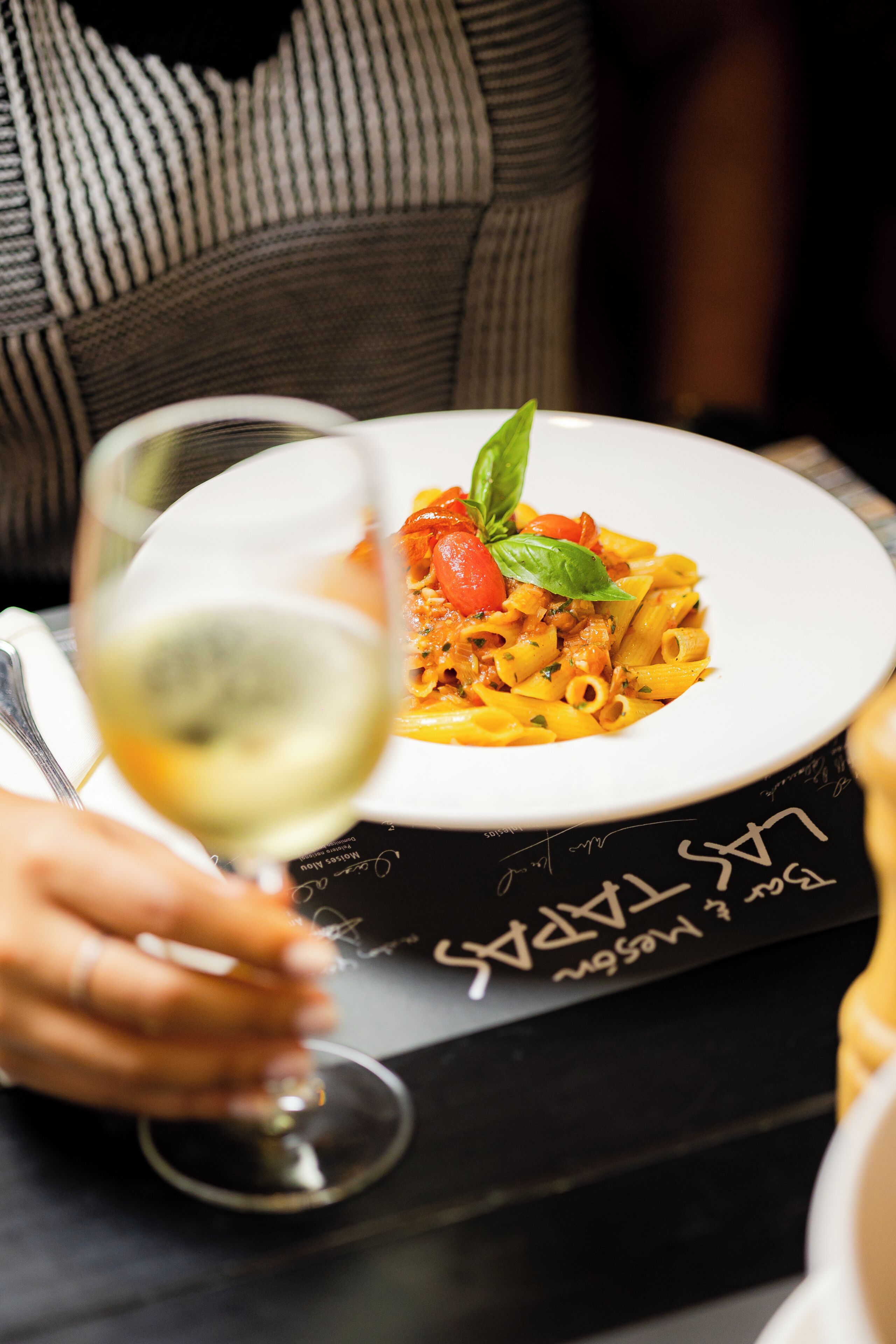 A person holding a glass of white wine, a plate of pasta with a basil leaf garnish, and a menu with text on a black tablecloth.