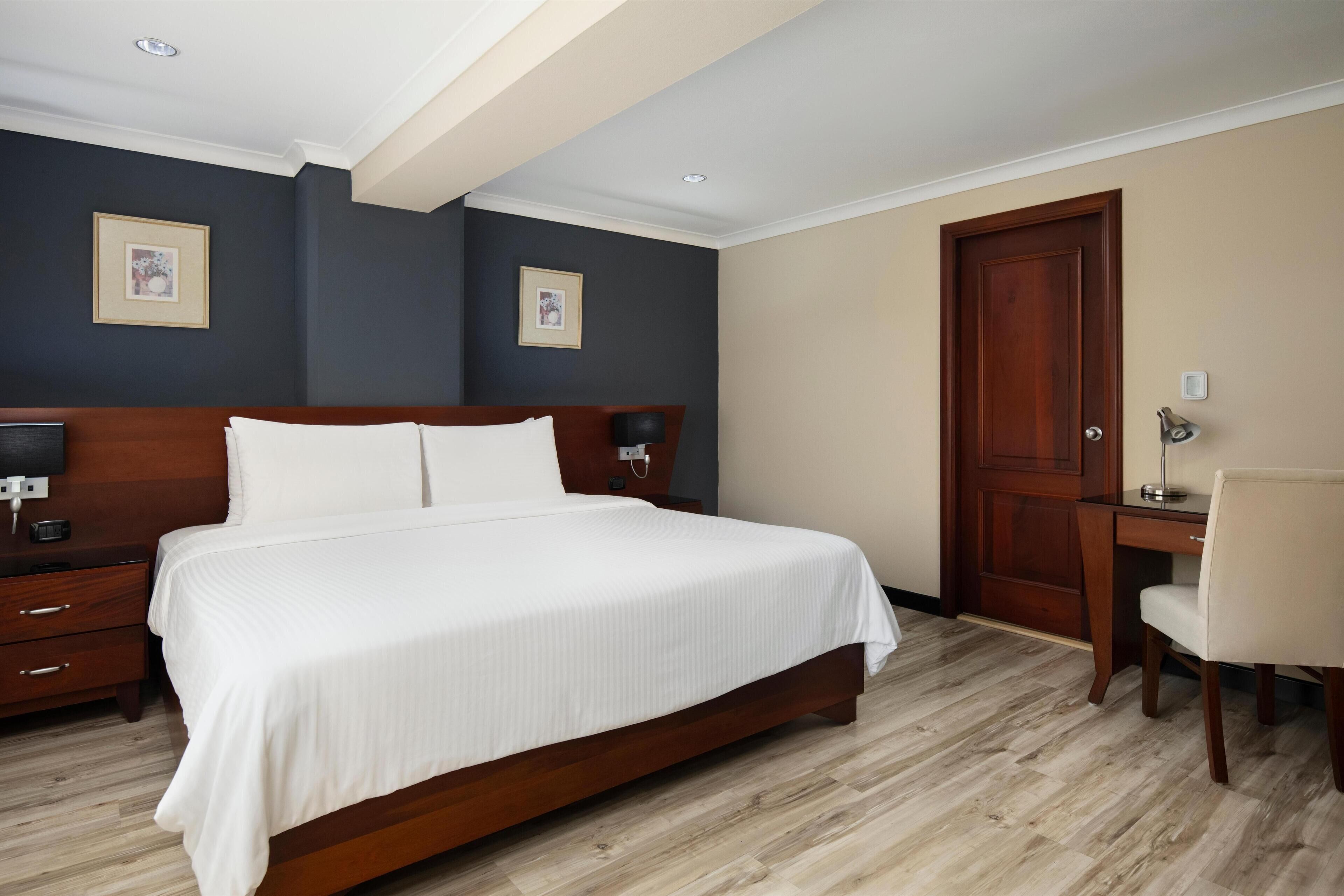 Habitación de hotel con una cama grande, cabecero de madera, un escritorio con silla y dos cuadros en las paredes.