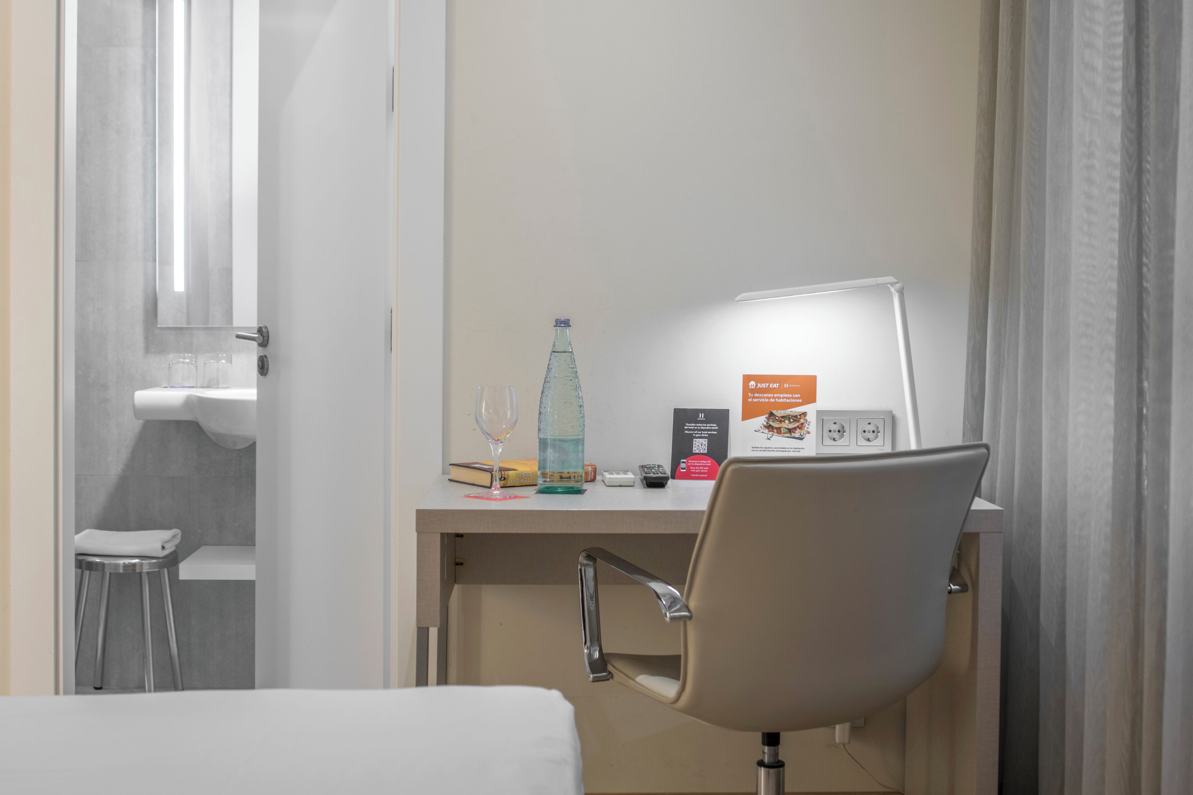 Une chambre d’hôtel avec un lit, un bureau avec une chaise, une bouteille, un verre, un livre, une télécommande et un panneau.