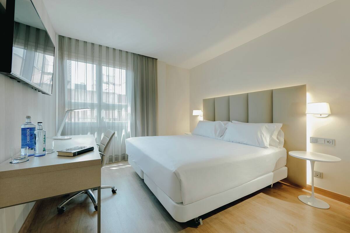 Ein Hotelzimmer mit Bett, Schreibtisch, Stuhl, Fernseher und einem Fenster mit Vorhängen.