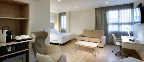 A modern hotel room with a bed, sofa, desk, and a kitchenette area.