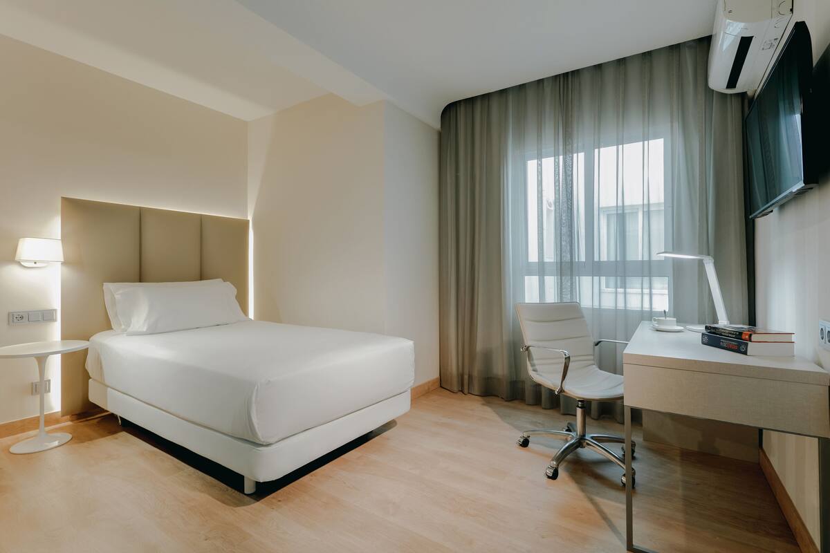 Ein modernes Hotelzimmer mit einem Bett, einem Schreibtisch, einem Sessel, einer Lampe und einem Fernseher.
