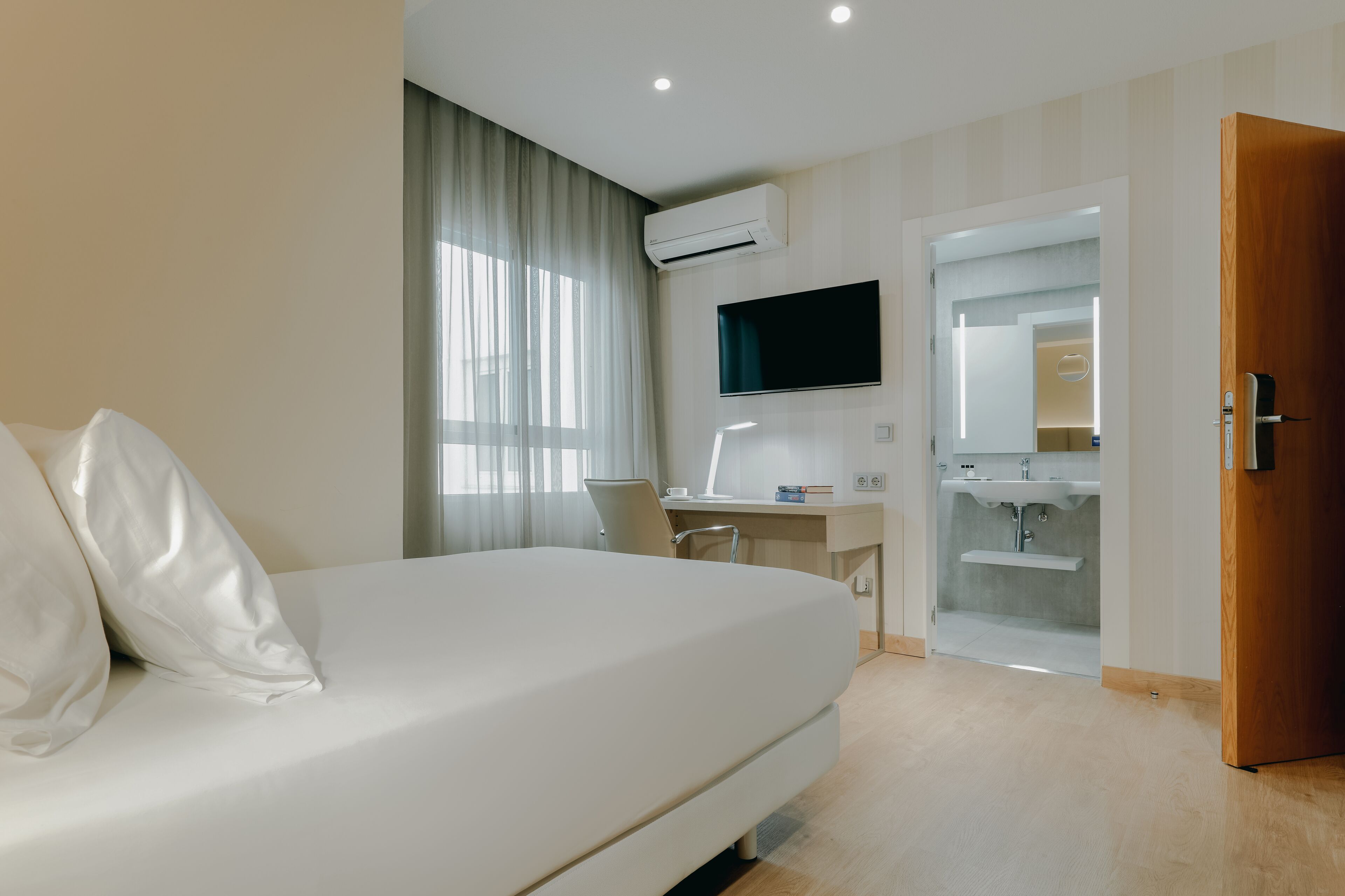 Habitación de hotel con cama, escritorio con computadora, televisión y baño visible a través de una puerta abierta.
