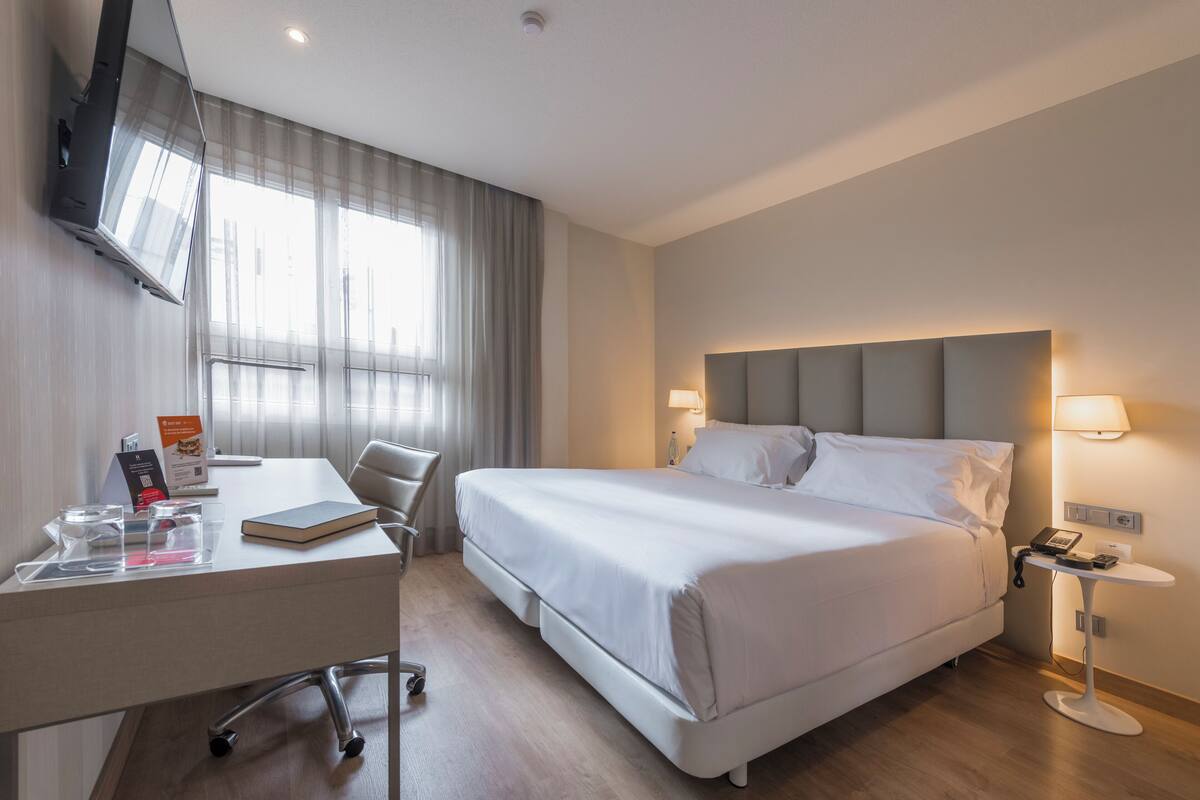 Ein Hotelzimmer mit Bett, Schreibtisch, Stuhl, Fernseher, Nachttischlampen und einem Fenster mit Vorhängen.