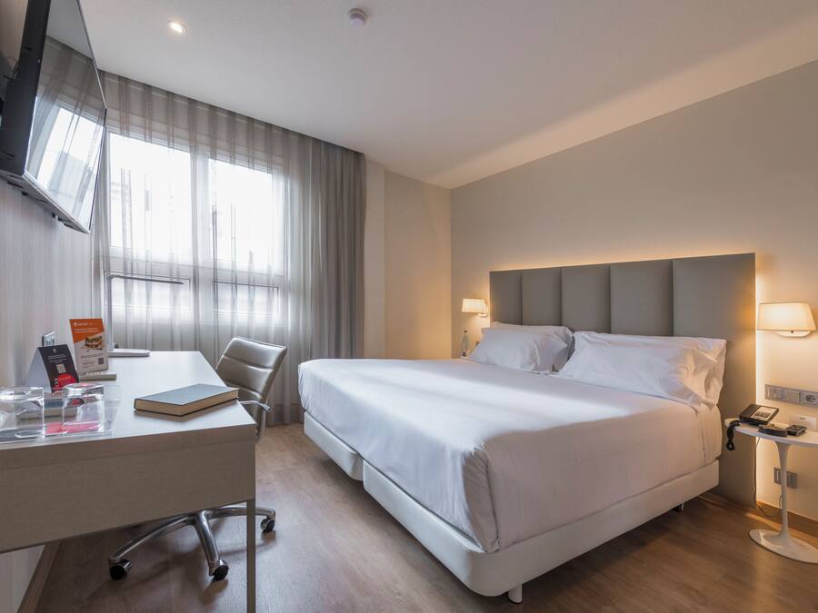A hotel room with a bed, bedside table, desk, chair, TV, and window with curtains.