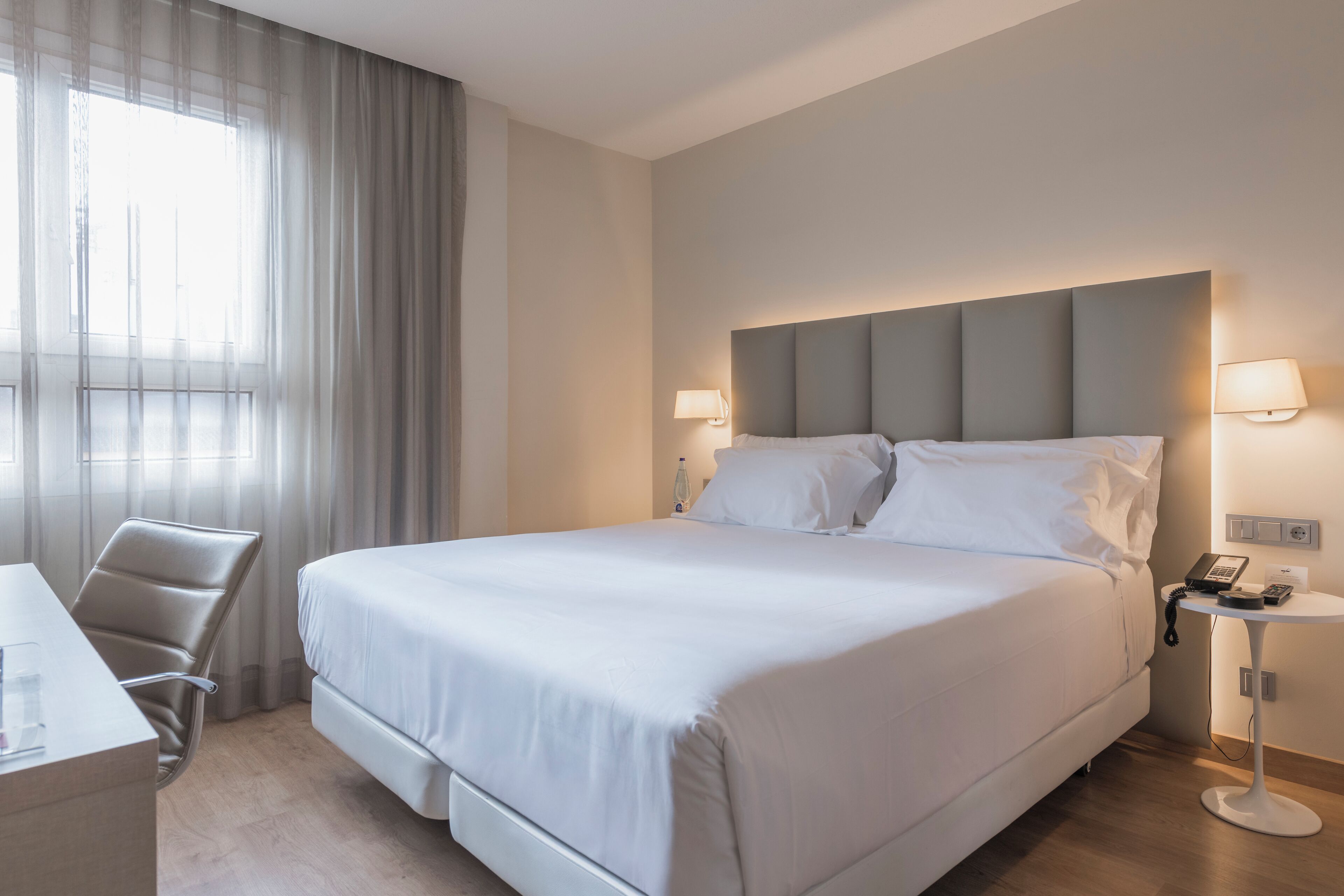 A hotel room with a large bed, a desk, a chair, and a bedside table with a lamp.
