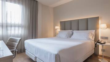 A hotel room with a large bed, a desk, a chair, and a bedside table with a lamp.