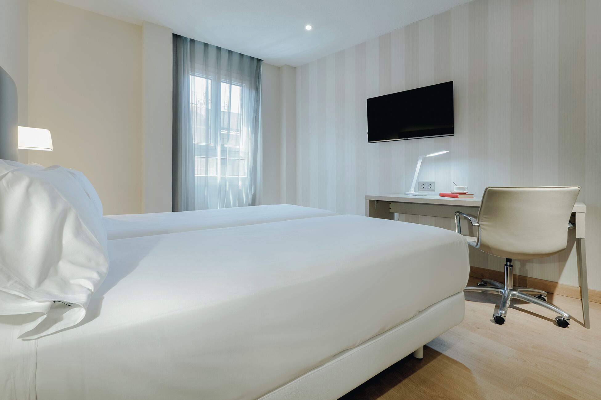 Una habitación de hotel con cama, escritorio con computadora, silla, televisor montado en la pared y ventana con cortinas.