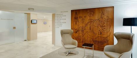 A modern lobby with two beige armchairs, a wooden wall with a cityscape mural, and a sign listing room names including 'Breakfast,' 'Meeting Room,' and 'Gym.'