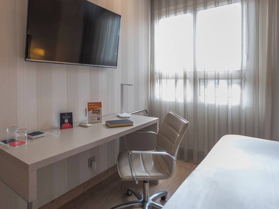 Ein Hotelzimmer mit Schreibtisch, Stuhl, Flachbildfernseher und einem großen Fenster mit Vorhängen.