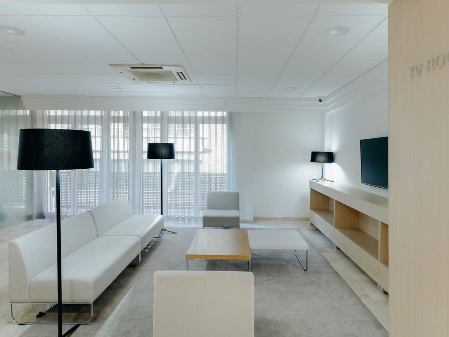 Un espacio de oficina moderno y bien iluminado con un sofá blanco, dos lámparas de pie negras, un televisor sobre un soporte de madera y un ventanal con persianas.
