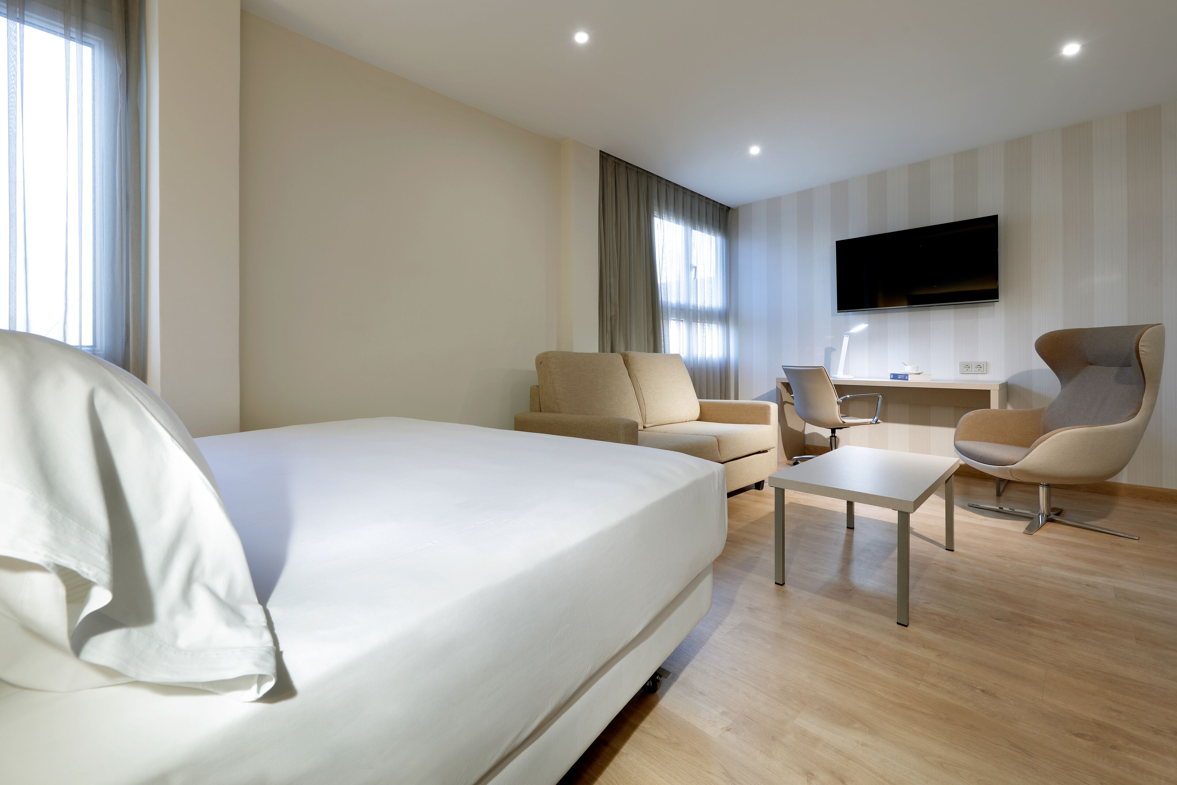 Une chambre d’hôtel moderne dotée d’un grand lit, de deux fauteuils, d’une petite table, d’un bureau et d’un téléviseur fixé au mur.
