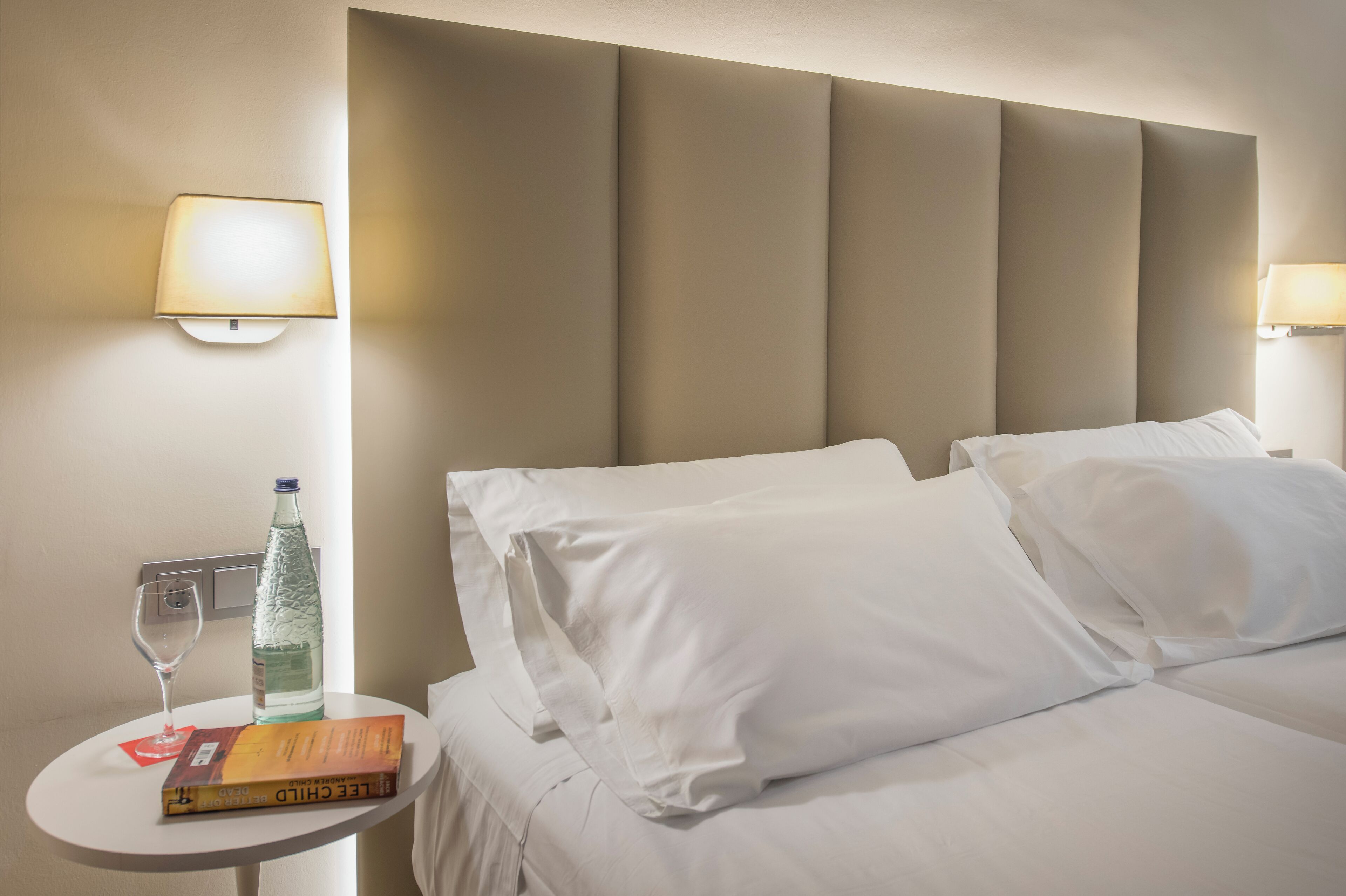 Habitación de hotel con cama, dos almohadas, mesita de noche con un vaso de agua, una botella y un libro.