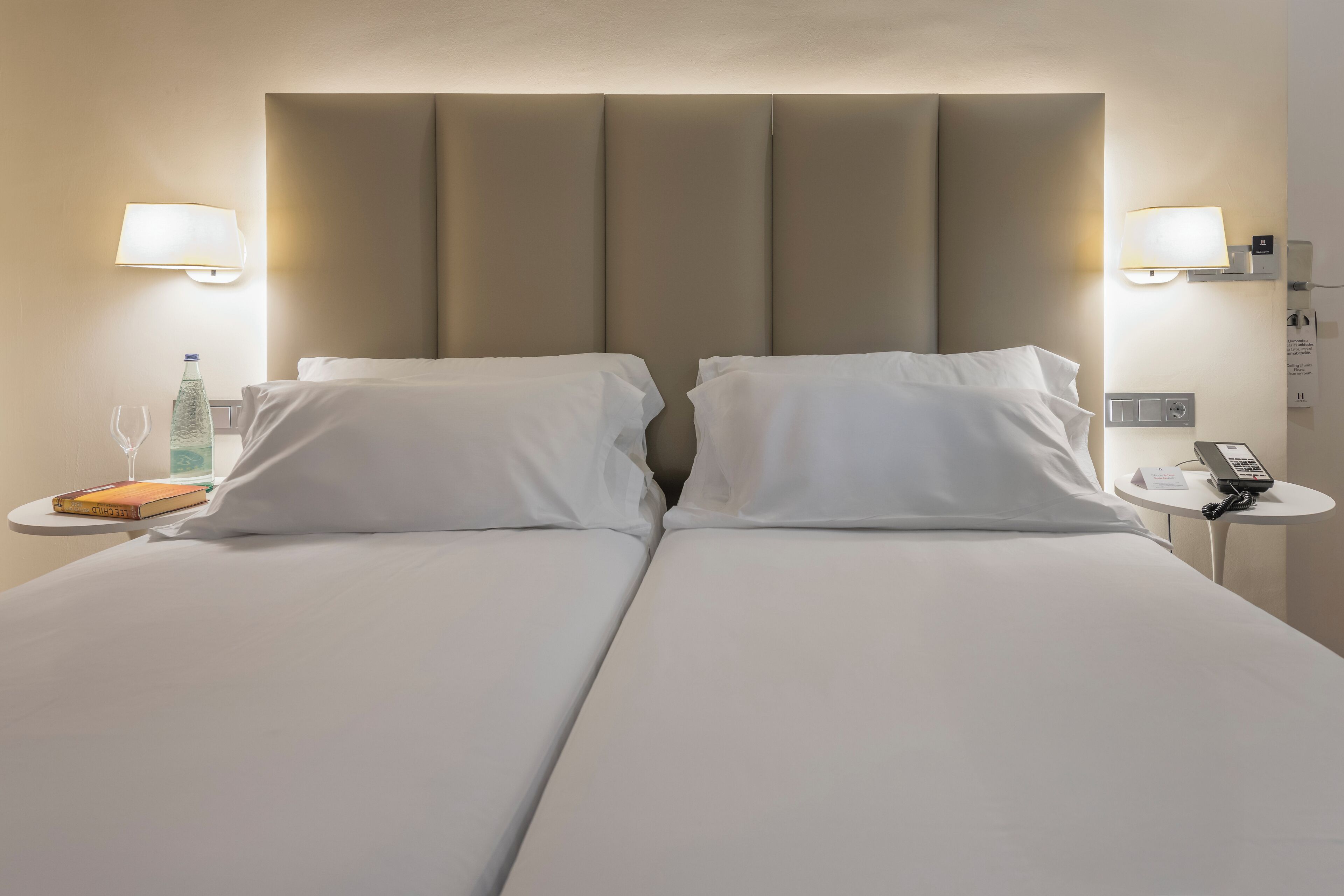 Habitación de hotel con una cama grande, dos mesitas de noche, una lámpara, una botella de agua y un libro.