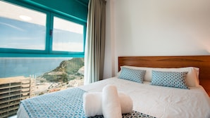 Une chambre d’hôtel avec un grand lit, deux coussins à motifs, une vue sur la mer et une fenêtre avec des rideaux.