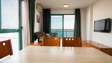 A hotel room with a large window, a dining table, two wooden chairs, a sofa, and a flat-screen TV.