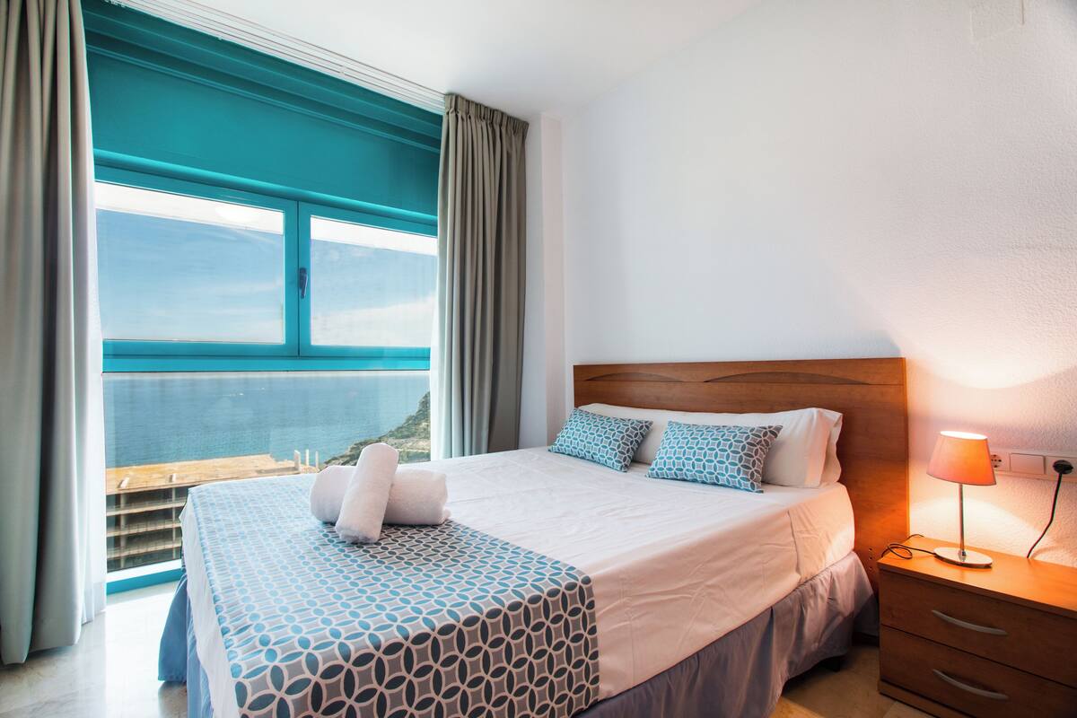 Una habitación de hotel con cama, mesita de noche, lámpara y ventana con cortinas.
