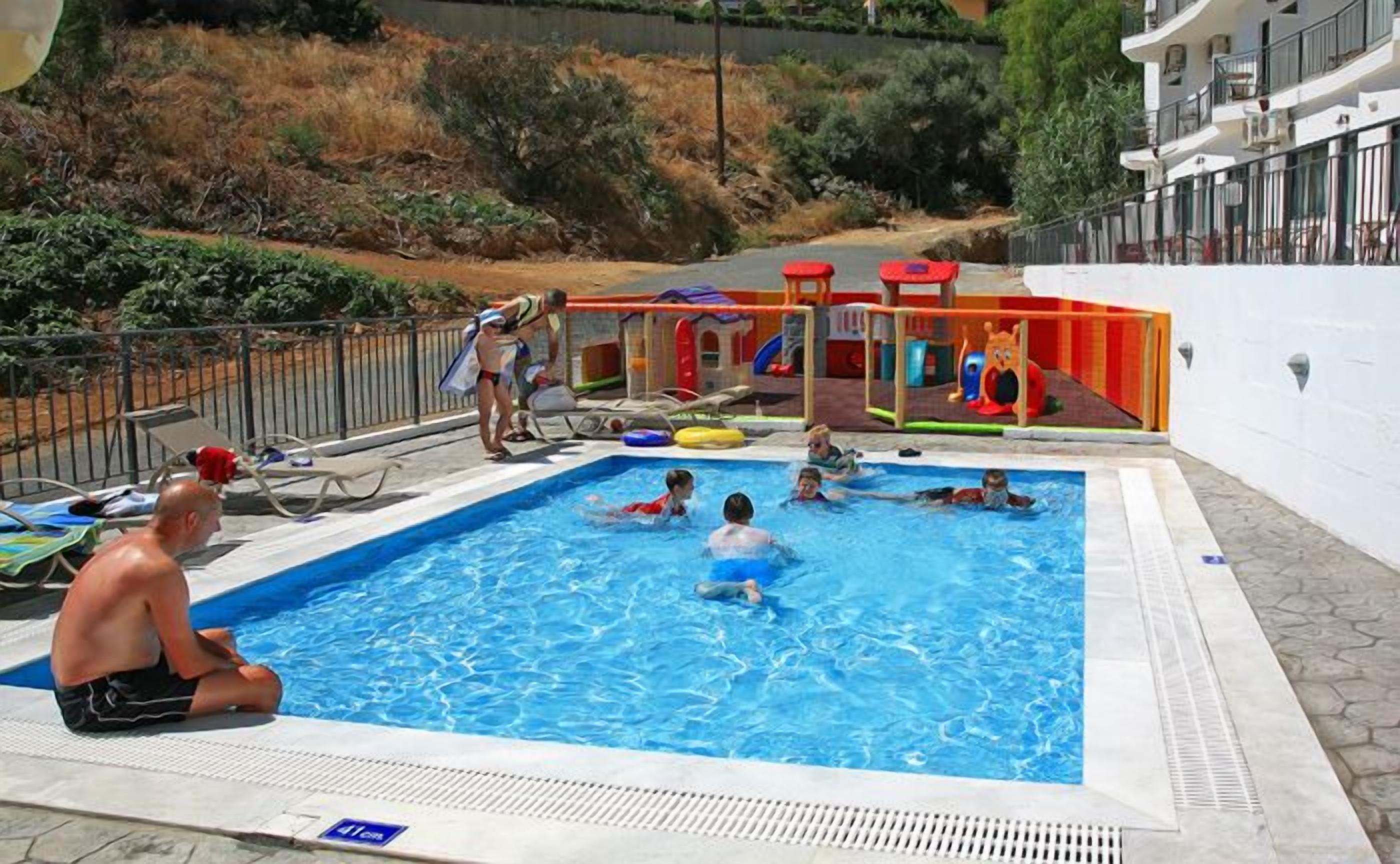 A swimming pool with children and adults in the water, surrounded by lounge chairs and a play area with colorful equipment.