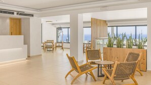 Una moderna hall d'hotel con una reception, un'area salotto con poltrone in vimini, ampie finestre con vista sull'oceano e un pannello in legno decorato con piante.