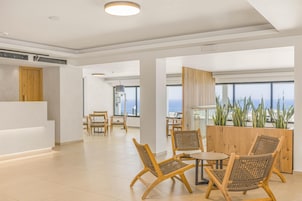 Un moderno vestíbulo de hotel con un mostrador de recepción, zona de estar con sillones de mimbre, ventanas amplias que ofrecen vistas al océano y un separador de madera con plantas.