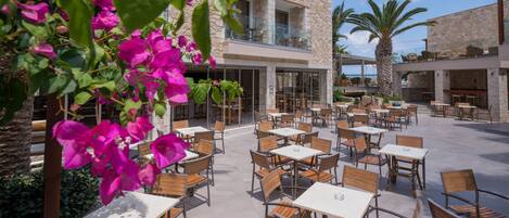 An outdoor patio area with multiple tables and chairs, a stone building with large windows, and a palm tree.