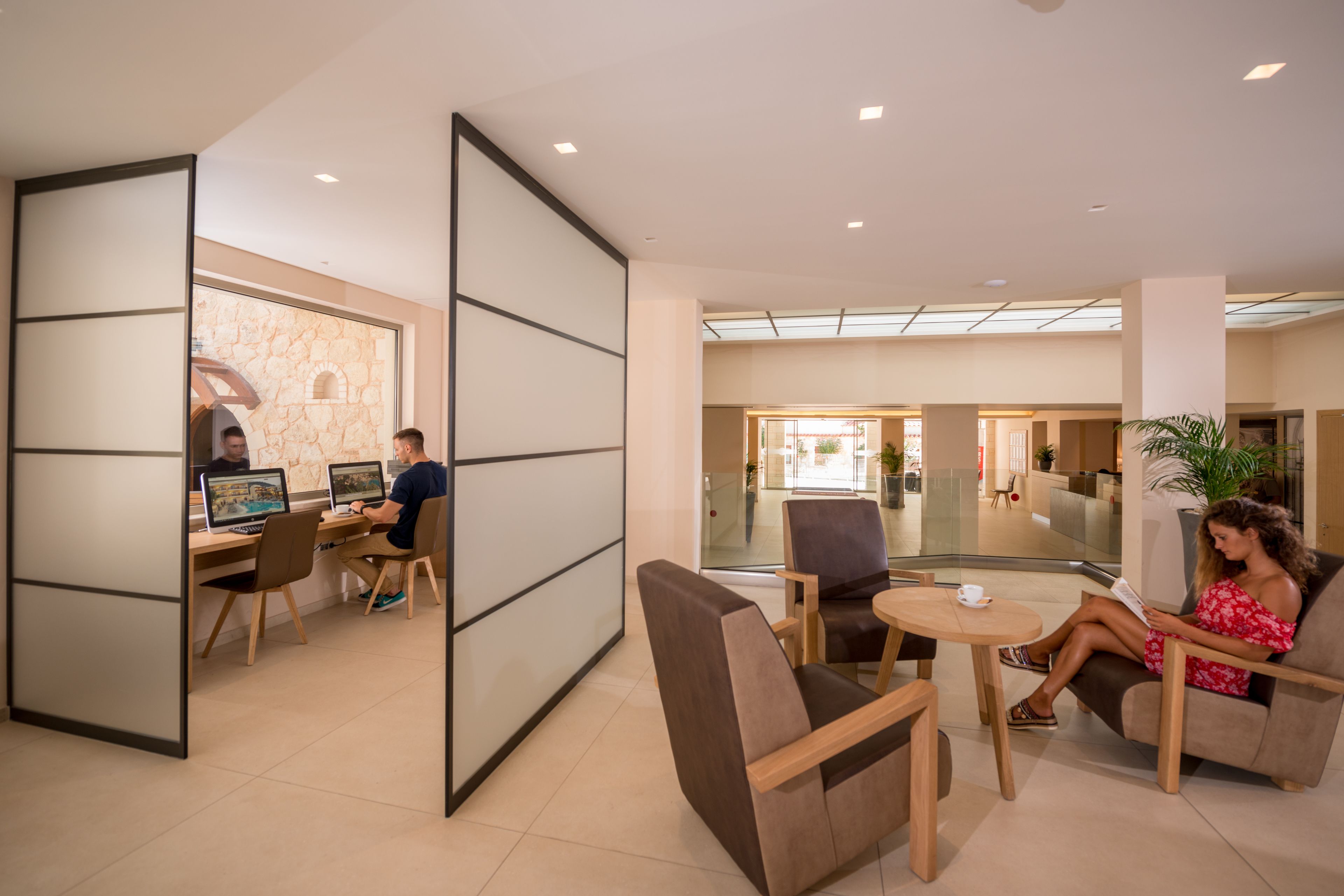 A modern lounge area with a glass partition, wooden furniture, and a view of the lobby.