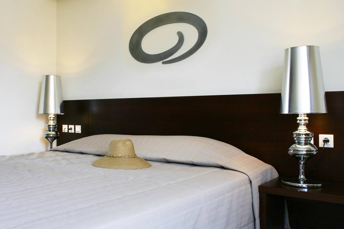 A neatly made bed with a hat on the pillow, two bedside lamps, and a wall-mounted abstract art piece.