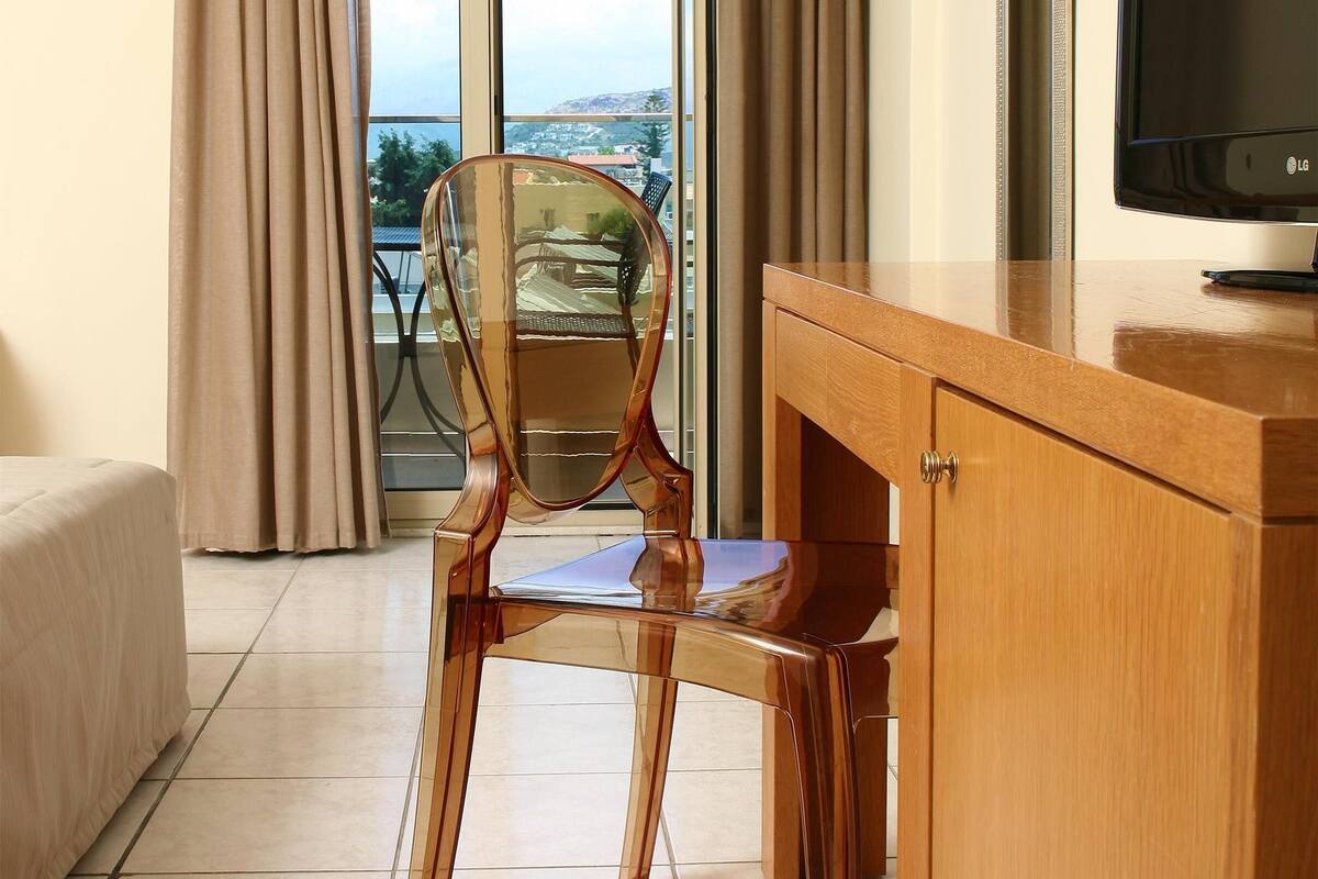 A hotel room with a wooden desk, a clear chair, a television, and a balcony with curtains.