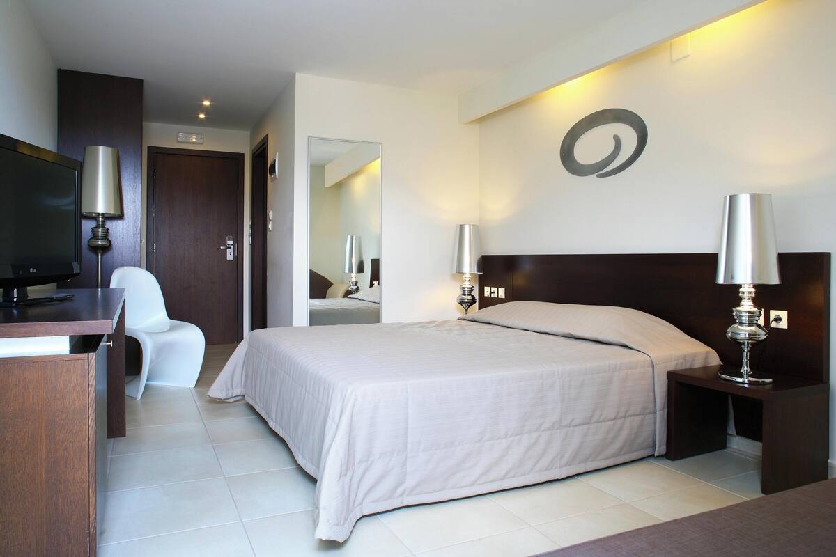 A hotel room with a bed, a desk, a chair, a TV, and two lamps.
