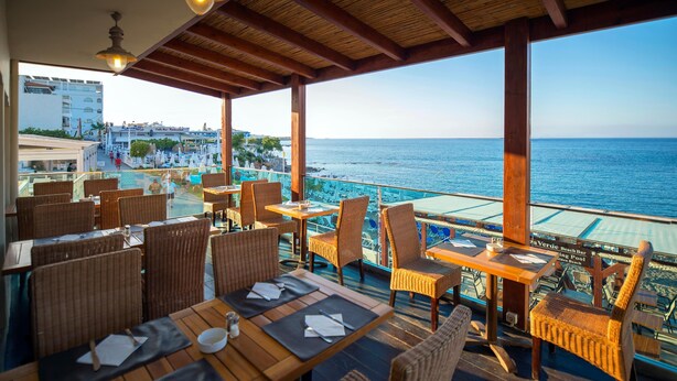 An outdoor dining area with wicker chairs and tables, overlooking a beach and the sea. - Golden Beach Hotel (Hersonissos)