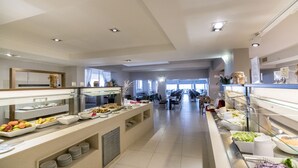 A spacious buffet area with multiple serving stations, a long counter, and a seating area in the background. - Golden Beach Hotel (Hersonissos)