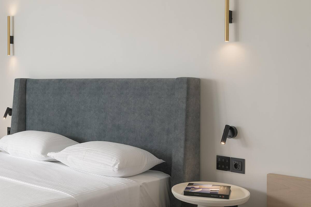 Quarto moderno com cabeceira cinza, roupa de cama branca e duas luminárias fixadas na parede.
